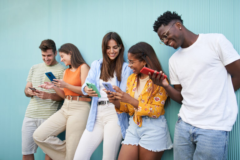Young smiling multicultural group of friends using cell phones. Students leaning against blue wall.