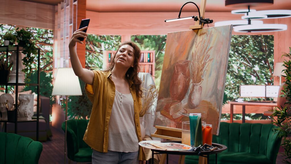 Woman taking a selfie shot with her phone of herself and her artwork behind her.