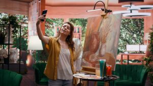 Woman taking a selfie shot with her phone of herself and her artwork behind her.