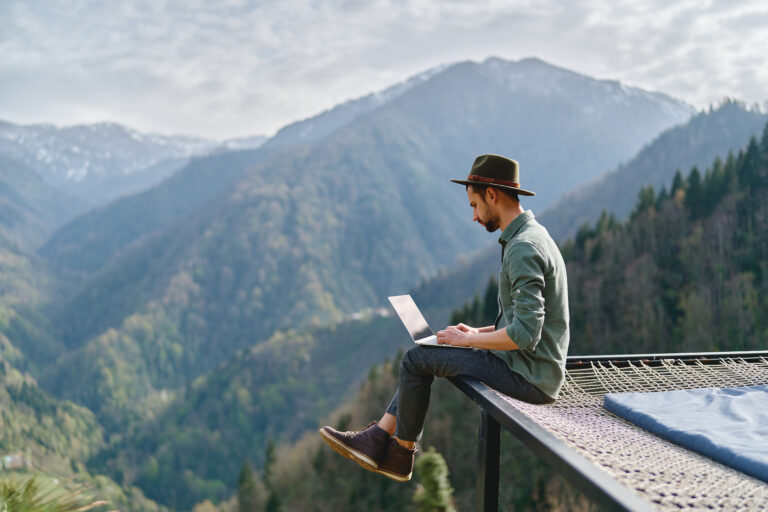 Young man with computer on his lap sitting at the edge of deck with mountain views